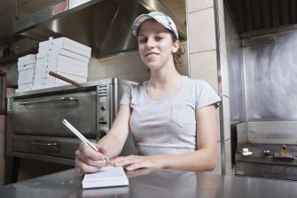 A young female worker taking orders in a restaurant kitchen setting.
