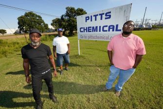 Three Black men stand in front of a sign for Pitts Chemical, highlighting their involvement in a lawsuit against Pitts Farm Partnership for alleged wage discrimination.