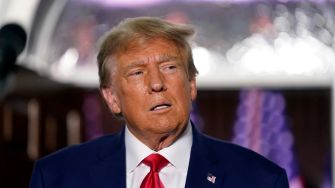 A close-up of a man with blond hair and a serious expression, wearing a dark suit and red tie.