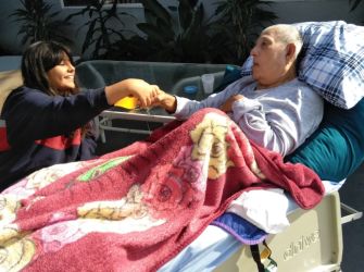 A woman and a man in a hospital bed share a moment together, with the man covered by a colorful blanket.
