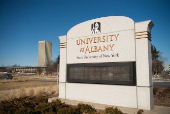 Signage for the University at Albany, State University of New York, with the campus buildings visible in the background.