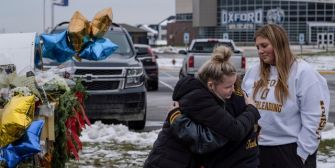 Two young girls embrace in front of a memorial at Oxford High School, surrounded by flowers and balloons, as the community mourns the aftermath of a recent school shooting.