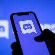A hand holding a smartphone displaying the Discord logo against a blue background. A hand holding a smartphone displaying the Discord logo against a blue background.
