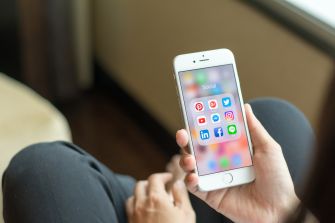 A person holding a smartphone displaying various social media app icons on the screen.