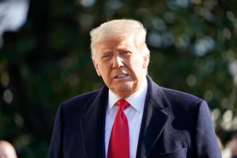 Donald Trump standing outdoors, wearing a suit and tie, with trees in the background.