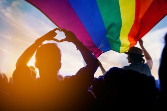 A silhouetted crowd raises their hands in support while a person waves a large rainbow flag, symbolizing Pride and LGBTQ+ rights.