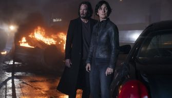 A scene from "The Matrix Resurrections" featuring two main characters standing in front of a burning vehicle.