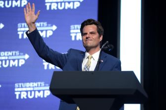 A man in a suit waves while standing at a podium with a "Students for Trump" backdrop.