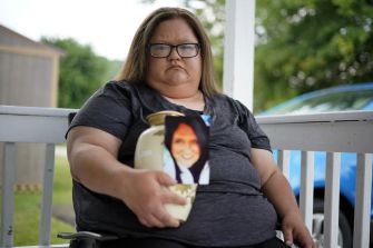 A woman sitting on a porch holding a urn and a photograph of a woman, expressing grief.