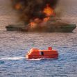 A boat on fire at sea with an orange lifeboat in the foreground. A boat on fire at sea with an orange lifeboat in the foreground.
