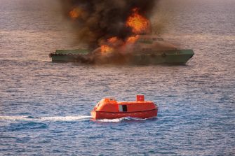 A boat on fire at sea with an orange lifeboat in the foreground.