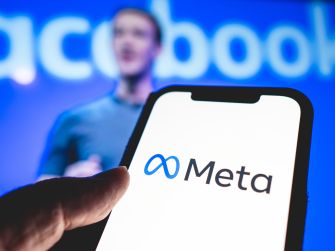 A person holding a smartphone displaying the Meta logo, with a blurred background of a man speaking at an event.