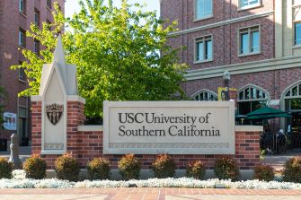Sign at the entrance of the University of Southern California (USC) with green trees and brick buildings in the background.