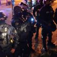 A group of police officers in riot gear, some with masks and helmets, standing in a dimly lit environment during a protest. A group of police officers in riot gear, some with masks and helmets, standing in a dimly lit environment during a protest.