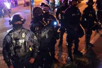 A group of police officers in riot gear, some with masks and helmets, standing in a dimly lit environment during a protest.
