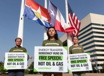 Three Greenpeace supporters holding signs that read "Energy Transfer: Democracy Runs on Free Speech, Not Oil & Gas" in front of flags and a building.