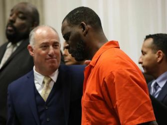 R. Kelly in an orange prison jumpsuit, accompanied by legal representatives in a courtroom setting.