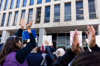 A crowd of protesters holding signs raises their arms in front of the U.S. Department of Education building.