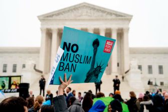 A protestor holding a sign that reads "No Muslim Ban" in front of the Supreme Court building, with a crowd gathered around.