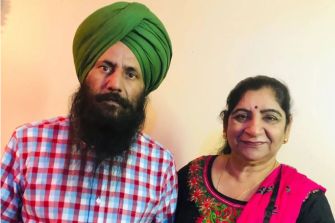 A Sikh man wearing a turban and a checked shirt stands next to a woman in traditional attire, both looking serious.