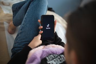 A person using a smartphone displaying the TikTok logo while relaxing on a bed.