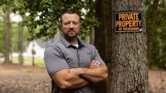 A man standing with his arms crossed next to a "Private Property No Trespassing" sign in a wooded area.