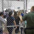 A border enforcement officer monitors a group of migrants lined up behind a fence in a detention facility. A border enforcement officer monitors a group of migrants lined up behind a fence in a detention facility.