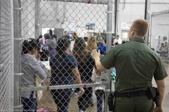 A border enforcement officer monitors a group of migrants lined up behind a fence in a detention facility.