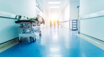 A hospital corridor with a medical stretcher in the foreground and healthcare workers in the background.