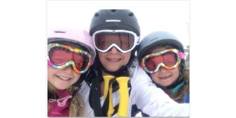 A woman and her two daughters, all wearing ski gear and helmets, smiling for a ski selfie.