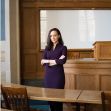 A law professor standing in a courtroom setting with arms crossed, wearing a purple dress. A law professor standing in a courtroom setting with arms crossed, wearing a purple dress.
