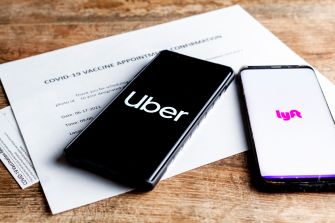Smartphones displaying the Uber and Lyft logos, positioned on a wooden surface next to a COVID-19 vaccine appointment confirmation document.