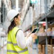 A worker in a safety vest and helmet uses a tablet while wearing a face mask in a warehouse environment. A worker in a safety vest and helmet uses a tablet while wearing a face mask in a warehouse environment.