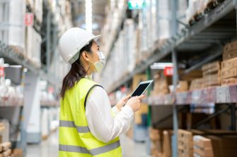 A worker in a safety vest and helmet uses a tablet while wearing a face mask in a warehouse environment.