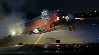 A red vehicle flipped over on a highway, with a police officer standing nearby.