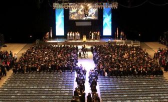 An aerial view of a graduation ceremony in a large venue, with graduates seated and a stage in the background.