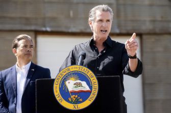 Governor Gavin Newsom speaking at a podium with the California state seal, addressing the media during a press conference.