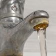 A close-up of a kitchen faucet with water flowing and visible mineral buildup and corrosion around the spout. A close-up of a kitchen faucet with water flowing and visible mineral buildup and corrosion around the spout.