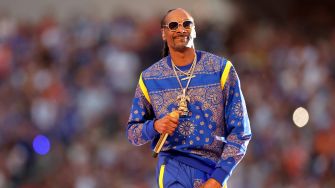 Snoop Dogg performing on stage at an event, dressed in a blue outfit with sunglasses and holding a microphone.