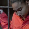 A young woman in an orange detention outfit looking down, standing near jail bars. A young woman in an orange detention outfit looking down, standing near jail bars.