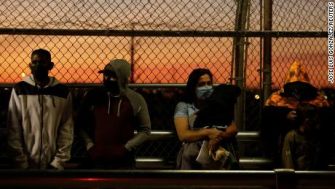 A group of migrants waiting on a bridge at sunset, with some wearing masks and hooded clothing, illustrating their uncertain journey and situation.