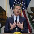 California Governor Gavin Newsom speaking at a press conference, with California flags in the background. California Governor Gavin Newsom speaking at a press conference, with California flags in the background.