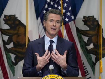 California Governor Gavin Newsom speaking at a press conference, with California flags in the background.