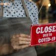 A person holding a red sign that reads "CLOSED DUE TO COVID-19," hanging it in a window. A person holding a red sign that reads "CLOSED DUE TO COVID-19," hanging it in a window.