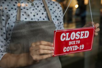 A person holding a red sign that reads "CLOSED DUE TO COVID-19," hanging it in a window.