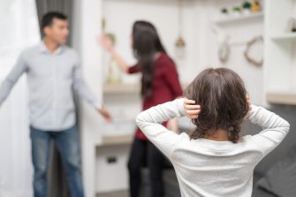 A young child covers her ears while two adults argue in the background, illustrating the impacts of domestic violence on family dynamics.