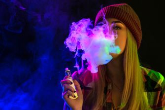A person exhaling colorful vapor from a vaping device in a dark setting.