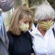 Allison Mack, wearing a black outfit and a mask, is assisted by a woman in a floral mask as they leave a courthouse surrounded by others. Allison Mack, wearing a black outfit and a mask, is assisted by a woman in a floral mask as they leave a courthouse surrounded by others.