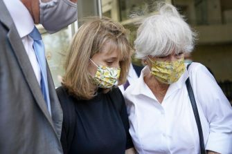 Allison Mack, wearing a black outfit and a mask, is assisted by a woman in a floral mask as they leave a courthouse surrounded by others.
