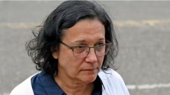 A woman with glasses and dark hair stands outside, appearing serious.